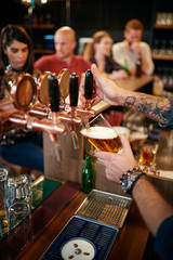 Tattooed caucasian barman pouring beer while standing in pub. Selective focus on hand.