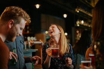 A small group of best friends standing at a pub, drinking beer, chatting and having fun. Focus on bartender wiping drinking glass. Nightlife.