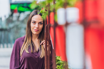 elegant and attractive woman with long straight hair dressed in shirt looking at camera and leaning next to some pots with red and white flowers