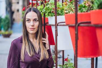 elegant and attractive woman with long straight hair dressed in shirt looking at camera and leaning next to some pots with red and white flowers