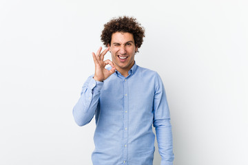 Young curly mature man wearing an elegant shirt winks an eye and holds an okay gesture with hand.