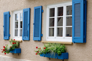 Two windows with white wooden frames, blue shutters and decorative flower boxes. Image of trendy decor, comfort, beautification. - Powered by Adobe