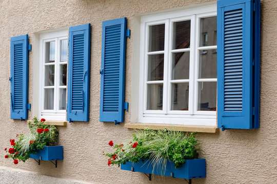 Two windows with white wooden frames, blue shutters and decorative flower boxes. Image of trendy decor, comfort, beautification.