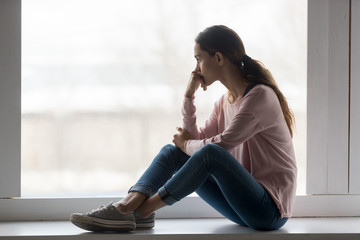 Frustrated young woman sitting on window sill at home.