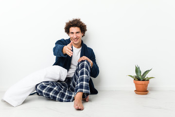 Mature man wearing pajama sitting on house floor stretching hand at camera in greeting gesture.