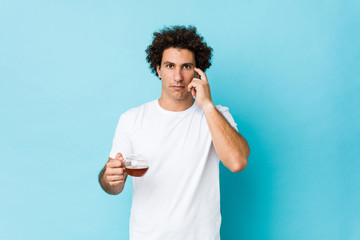 Young caucasian curly man holding a tea cup pointing his temple with finger, thinking, focused on a task.