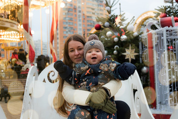 A happy little boy in a warm jacket and a knitted hat rides a carousel horse during a family trip to the Christmas market. A little boy with his mother on the carousel. 