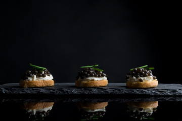 Caviar, three snacks of black caviar on bread and stone plate isolated on black background with reflections, copy space - obrazy, fototapety, plakaty