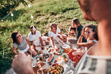 A happy group of teenage friends sitting on blanket in the park drinking beer and eating their lunch