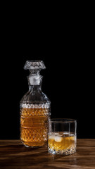 Glass of whiskey with ice and a square decanter isolated on black background on wooden table. Alcohol concept