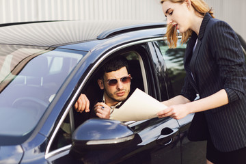 A young man rents a car. Employee of the dealer center shows documents near the car