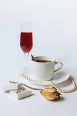 A still life with a white ceramic cup of coffee, a faded rose, a coral necklace, a little white box with the silver earrings, and the glass of champagne isolated on a white background