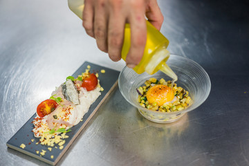 Chef hands pouring olive oil on a modern kitchen plate accompanied by a table with seafood salad next to it