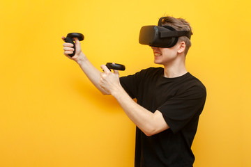 guy in VR glasses on a yellow background plays a virtual shooter, an emotional gamer shoots a game with joysticks and screams
