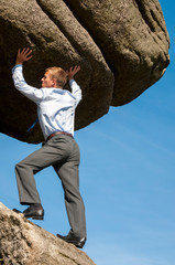 Strong businessman struggling to lift massive boulder into blue sky copy space