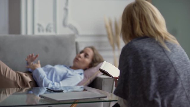 Rack focus of calm young woman gesturing lying on sofa and explaining problem to counselor with clipboard in hand nodding while listening to client