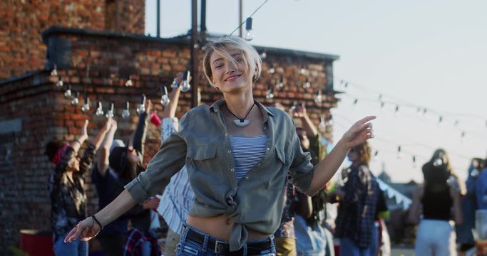 Young stylish and attractive Caucasian woman with short blond hair dancing in front of the camera at the rooftop party. Outdoor.