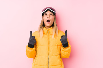 Young caucasian woman wearing a ski clothes in a pink background pointing upside with opened mouth.
