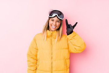 Young caucasian woman wearing a ski clothes in a pink background showing a horns gesture as a revolution concept.