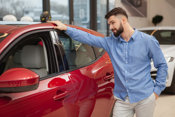 Fototapeta na wymiar Handsome bearded man examining red automobile at car dealership. Male customer buying new auto