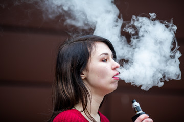 Vape teenager. Young pretty white caucasian girl in a red sweater smoking an electronic cigarette opposite modern brown background on the street in the winter. Deadly bad habit.