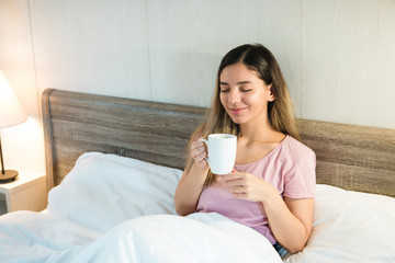 smiling beautiful woman with eyes closed holding a cup while lying in bed with white bedding