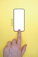 Abstract year 2020 of wooden letters next to a hand push to smartphone on a yellow background. Top view. Mock up. Copy space.