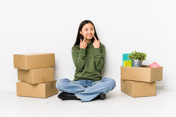 Young chinese woman moving to a new home smiles, pointing fingers at mouth.
