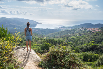 Young woman admiring panorama from hill overseeing bay in Tuscany wearing denim dungaree