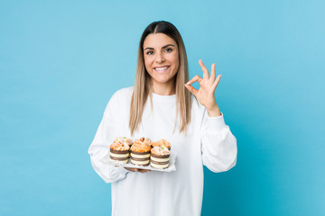 Young caucasian woman holding a sweets cake cheerful and confident showing ok gesture.