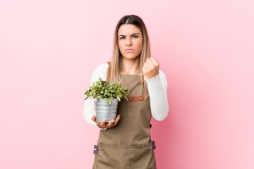 Young gardener woman holding a plant showing fist to camera, aggressive facial expression.