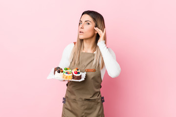 Young baker woman holding sweets trying to listening a gossip.