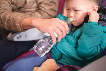 Asian 2 - 3 years old toddler boy child holding his aching ear and drinking water from bottle during flight on airplane. Little kid feeling earache on airplane, Flying with children, Selective focus