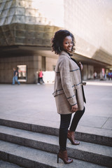 Obraz premium Beautiful african american young woman with afro and large hoop earrings in a stylish coat, smiling. Urban street portrait.