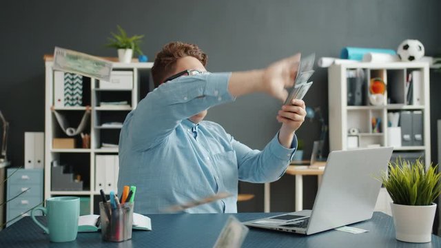 Successful businessman young man is throwing money having fun at desk in office alone enjoying wealth. Rich people, lifestyle and business concept.