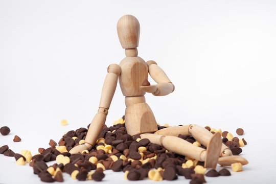 wooden jointed mannequin sitting in pile of dark chocolate chips white chocolate and milk chocolate on solid white background