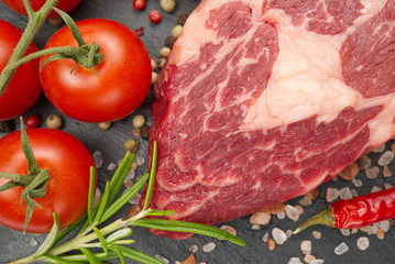 Raw rib eye steak with spices and vegetables. Ingredients for restaurant meal. Fresh meat, salt, rosemary, thyme, chilli, cherry tomatoes, garlic on black stone. Food background.