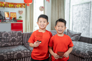 two asian chinese boys smile wearing red shirts holding angpau standing at front sofa