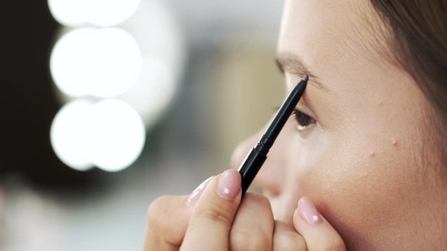 A makeup artist draws the eyebrows. She is working. A lady has brown eyes.