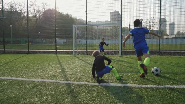 Children's soccer team playing football outdoors, forward bypassing back player and scoring goal, confused goalkeeper missing second goal from other forward. Happy children rejoicing in goals scored