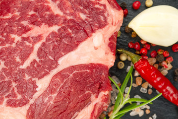 Raw rib eye steak with spices and vegetables. Ingredients for restaurant meal. Fresh meat, salt, rosemary, thyme, chilli, cherry tomatoes, garlic on black stone. Food background.