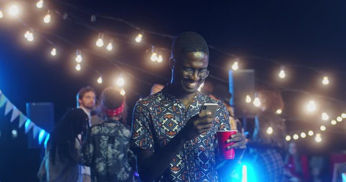 Young happy smiled African American guy in glasses standing at the party outside at night and using his smartphone, tapping and chatting.