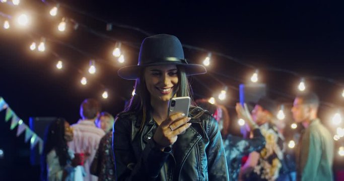 Attractive young Caucasian girl in a hat taping and texting on the smartphone at the party late in the evening. Outdoors.