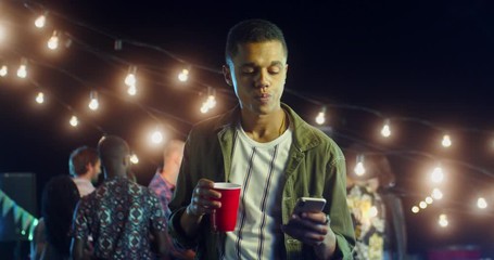 Young good looking stylish moulatto guy sipping a drink and tapping or texting on the smartphone while being at the aprty at night.