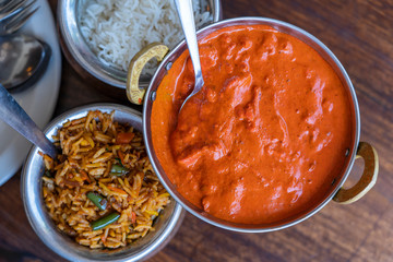 Navratan saffron biryani with fresh vegetables, spices and basmati rice in a metal pot and pieces of chicken in red curry sauce on wooden background, close up. Indian cuisine