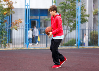 Obraz premium Cute boy in red t shirt plays basketball on city playground. Active teen enjoying outdoor game with orange ball. Hobby, active lifestyle, sport for kids.