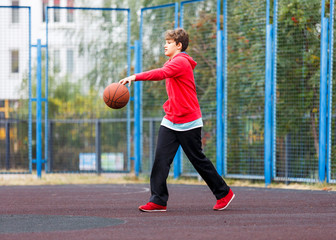 Cute boy in red t shirt plays basketball on city playground. Active teen enjoying outdoor game with orange ball. Hobby, active lifestyle, sport for kids.