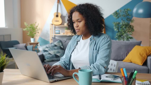 Slow motion of African American girl using laptop in apartment smiling typing enjoying freelance work sitting at desk indoors . Electronic devices and freelance work concept.