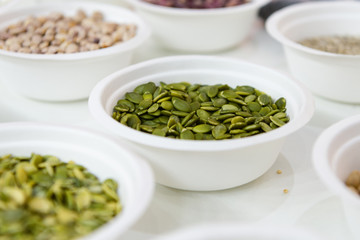 Peeled pumpkin seeds lie in a white plastic plate. Around in the same plates are various seeds, nuts and legumes.