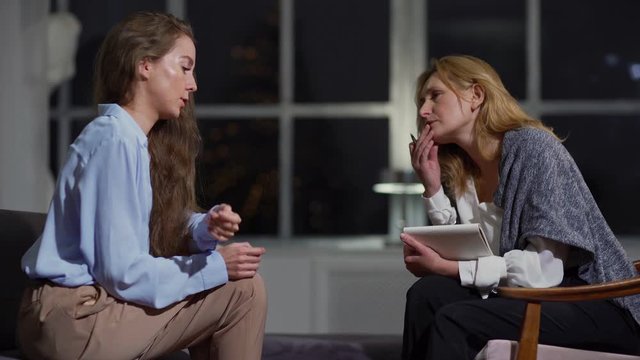 Stressed young woman talking emotionally about her problems to middle aged female therapist listening attentively, taking notes in notepad and giving advice at evening session
